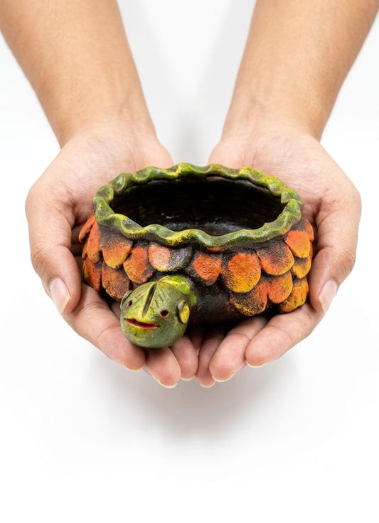 A small decorative turtle-shaped planter held in two hands against a plain white background. The planter has a dark body, orange and yellow scalloped shell, and a green head and rim.